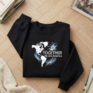 Puede incluir: Sudadera negra con un gr&aacute;fico blanco de un mapa del mundo y detalles en azul. El texto "TOGETHER We Are America" est&aacute; debajo del mapa. La sudadera est&aacute; hecha de un material suave.