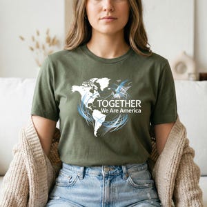 Puede incluir: Camiseta verde oliva con un dise&ntilde;o de mapa del mundo blanco, detalles en azul y el texto "TOGETHER We Are America." La camiseta est&aacute; hecha de un material suave y es adecuada para uso casual.