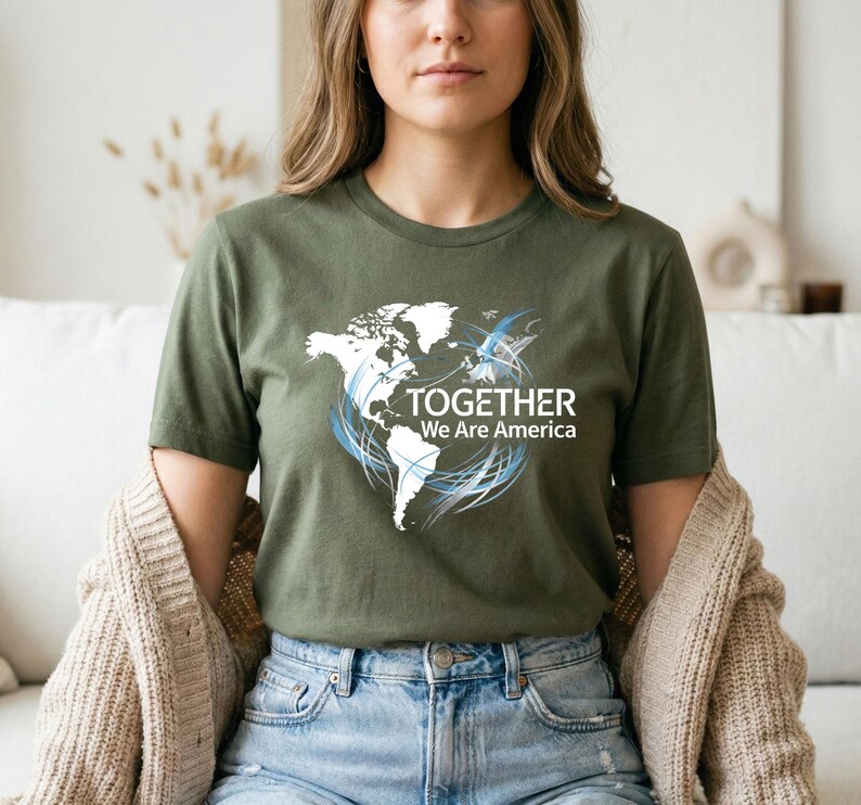 Puede incluir: Camiseta verde oliva con un dise&ntilde;o de mapa del mundo blanco, detalles en azul y el texto "TOGETHER We Are America." La camiseta est&aacute; hecha de un material suave y es adecuada para uso casual.