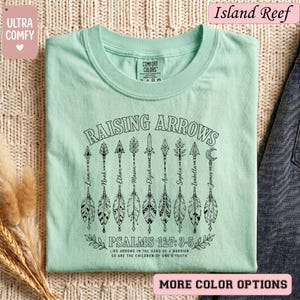 Puede incluir: Una camiseta de color verde menta claro con un gráfico negro de nueve flechas con nombres escritos en los astiles. El texto dice RAISING ARROWS, PSALMS 127:3-5 y LIKE ARROWS IN THE HAND OF A WARRIOR, SO ARE THE CHILDREN OF ONE'S YOUTH.