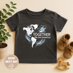 Puede incluir: Camiseta gris oscuro para ni&ntilde;os peque&ntilde;os con un gr&aacute;fico blanco y azul de un mapa del mundo y el texto "TOGETHER We Are America". La camiseta se muestra con zapatos marrones y un caballito de madera.