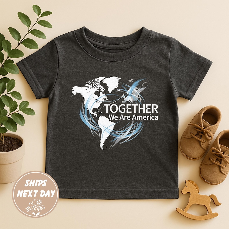 Puede incluir: Camiseta gris oscuro para ni&ntilde;os peque&ntilde;os con un gr&aacute;fico blanco y azul de un mapa del mundo y el texto "TOGETHER We Are America". La camiseta se muestra con zapatos marrones y un caballito de madera.