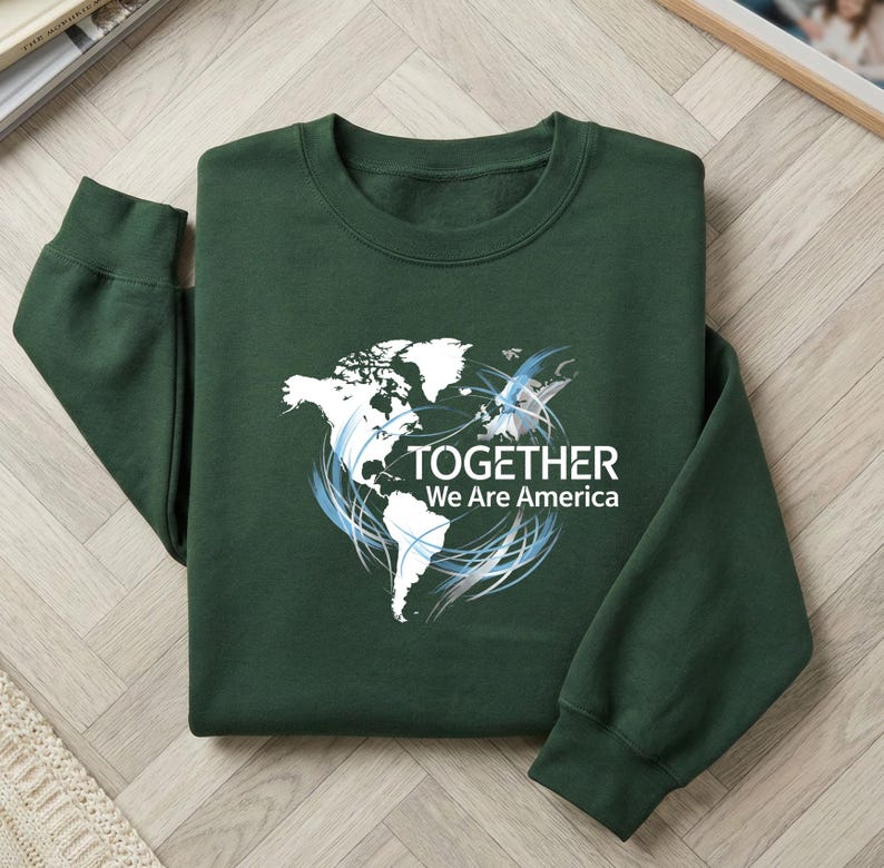 Puede incluir: Sudadera verde oscuro con un gr&aacute;fico de mapa del mundo blanco y el texto "TOGETHER We Are America". El dise&ntilde;o incluye remolinos azules y plateados. La sudadera est&aacute; hecha de un material suave.