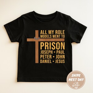 May include: Black t-shirt with a wooden cross graphic and the text "ALL MY ROLE MODELS WENT TO PRISON" with a list of names. A round sticker with the text "SHIPS NEXT DAY" is on the bottom right.