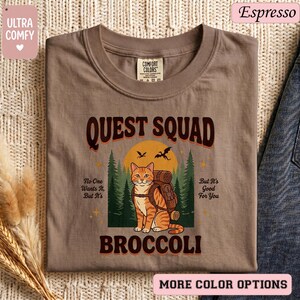 May include: Espresso-colored t-shirt with the text "QUEST SQUAD BROCCOLI" and a graphic of a cat with a backpack in front of a sunset and trees. The shirt also has the text "No One Wants It, But It's" and "But It's Good For You".