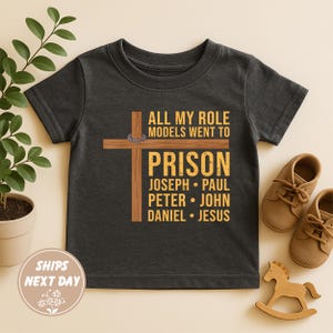 May include: Dark gray toddler t-shirt with the text "ALL MY ROLE MODELS WENT TO PRISON" and a cross graphic. The names Joseph, Paul, Peter, John, Daniel, and Jesus are listed below. Brown shoes and a wooden rocking horse are in the image.