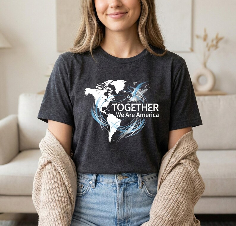 Puede incluir: Camiseta gris oscuro con un gr&aacute;fico de mapa del mundo blanco y el texto "TOGETHER We Are America". El dise&ntilde;o incluye remolinos azules alrededor del mapa. La camiseta est&aacute; hecha de un material suave.
