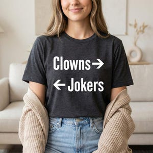 May include: Dark gray t-shirt with the text "Clowns -> Jokers" in white. The shirt is a crew neck style and is made of a soft material. The shirt is paired with blue jeans.