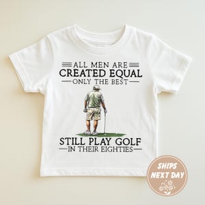 Op de afbeelding: Wit t-shirt met de tekst "ALL MEN ARE CREATED EQUAL - ONLY THE BEST - STILL PLAY GOLF - IN THEIR EIGHTIES." Het t-shirt heeft een afbeelding van een golfer. Het t-shirt heeft korte mouwen.