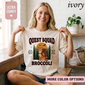 May include: Ivory-colored t-shirt with the text "QUEST SQUAD" in a retro font. The graphic features an orange tabby cat with a backpack, set against a forest and sunset backdrop. The text "BROCCOLI" is below the cat.