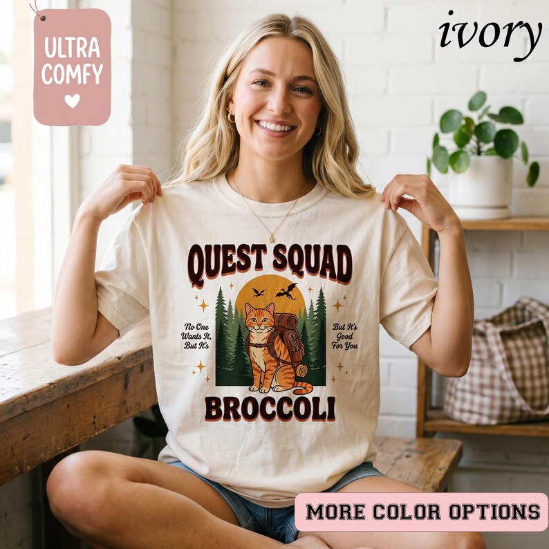May include: Ivory-colored t-shirt with the text "QUEST SQUAD" in a retro font. The graphic features an orange tabby cat with a backpack, set against a forest and sunset backdrop. The text "BROCCOLI" is below the cat.