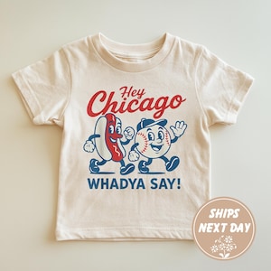 Könnte beinhalten: Cremefarbenes T-Shirt mit einem Cartoon-Hotdog- und Baseball-Charakter. Der Text "Hey Chicago WHADYA SAY!" ist in Rot und Blau gehalten. Der Baseball-Charakter trägt eine blaue Kappe. Das Shirt hat einen Rundhalsausschnitt und kurze Ärmel.