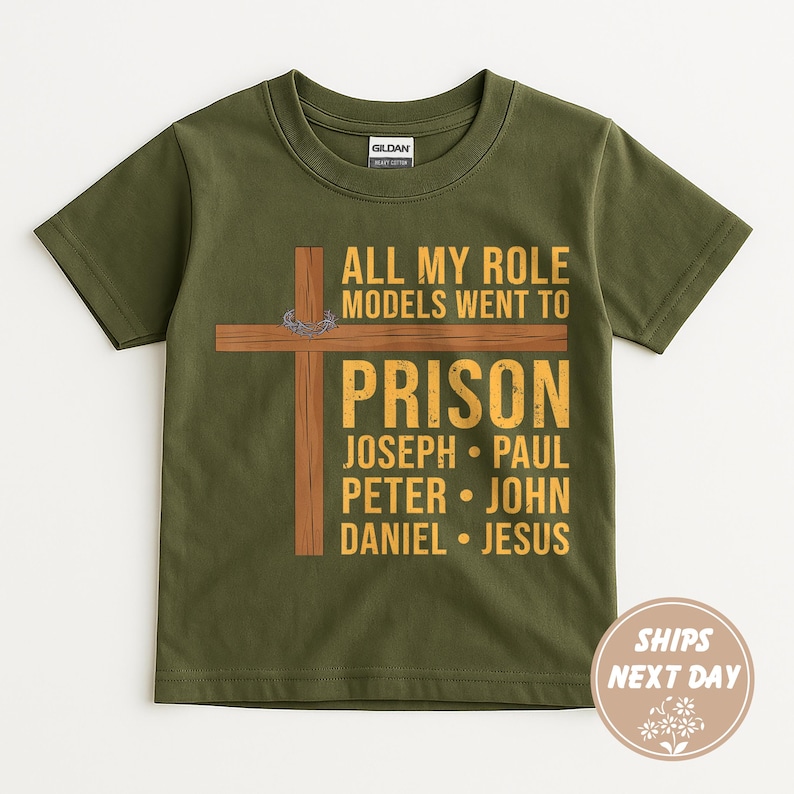 May include: Olive green t-shirt with a graphic featuring a wooden cross and the text "ALL MY ROLE MODELS WENT TO PRISON" followed by names. The shirt has a crew neck and short sleeves. The words "SHIPS NEXT DAY" are also visible.