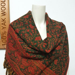 May include: A green and red paisley patterned shawl with a fringed edge. The shawl is draped over a mannequin, showcasing the intricate floral and paisley design. The text "100% YAK WOOL" is visible in the background.