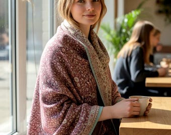 May include: A burgundy shawl with a floral pattern and a light blue border. The shawl is draped over the shoulders. The shawl has a fringed edge. The person is sitting at a wooden table.