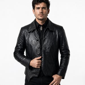 May include: A black leather jacket featuring a pointed collar, long sleeves, and a front closure with three black buttons. The jacket has curved seam detailing on the chest and a slightly asymmetrical hemline, worn over a black shirt with a bolo tie.
