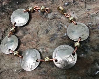 May include: A bracelet made with copper and gold chain links and five silver buffalo nickel coins. The coins are connected with the chain links and gold beads.