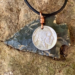 May include: A handcrafted necklace featuring a silver 1937 Buffalo nickel pendant, suspended from a black leather cord. The pendant is attached with copper wire to a dark gray arrowhead-shaped stone. The Fun Co. logo is visible in the lower left corner.