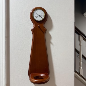 May include: A wooden barometer with a circular gauge at the top and a small shelf at the bottom. The barometer is mounted on a wall and has a dark brown finish. The gauge has a white face with black markings and a gold-colored frame.
