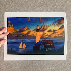 May include: A colorful painting of a lighthouse at sunset. The sky is filled with orange and blue hues, and a sailboat is visible on the water. A house with a red roof sits next to the lighthouse.