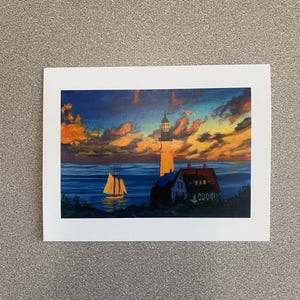 May include: A scenic print of a lighthouse at sunset. The artwork features a white lighthouse with a black top, a sailboat on the water, and a colorful sky with orange and blue hues. The lighthouse is next to a red-roofed building.