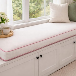 Op de afbeelding: Een wit vensterbankkussen met een roze rand, geplaatst op een witte kast met zwarte knoppen. De vensterbank staat naast een raam met witte gordijnen. Er ligt een klein tapijt op de vloer.