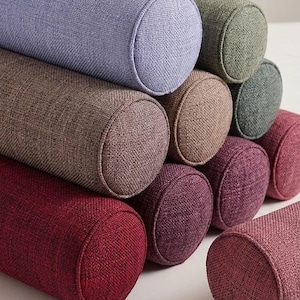 May include: A selection of cylindrical bolster cushions in shades of lavender, olive green, and burgundy. The cushions have a textured fabric and rounded ends, arranged on a white surface.