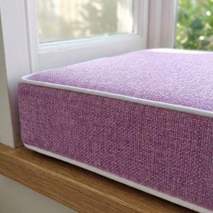 Op de afbeelding: Een rechthoekig lavendelkleurig vensterbankkussen met een witte bies. Het kussen is gemaakt van een textuurstof en ligt op een houten vensterbank. Het raam is zichtbaar op de achtergrond.