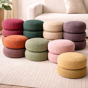 Puede incluir: Una colección de cojines redondos para el suelo en varios colores, incluyendo burdeos, verde, beige, morado, naranja, verde claro, rosa y amarillo. Los cojines están apilados en pares y colocados sobre una alfombra de color neutro.