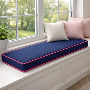 Può includere: Un cuscino blu navy per la seduta della finestra con bordatura rosa. Il cuscino è rettangolare e si trova su una seduta bianca. Diversi cuscini bianchi e rosa sono sulla seduta.
