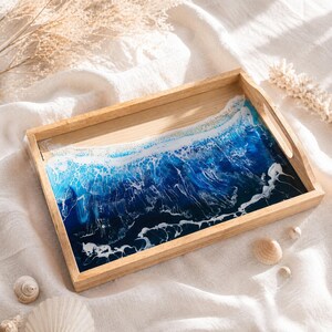 May include: Wooden serving tray with a resin ocean wave design. The tray has a light wood frame and handles. The ocean design features shades of blue and white, creating a realistic wave effect. Decorative seashells are scattered around the tray.