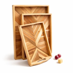 May include: Three wooden serving trays with a chevron pattern. The trays are made of light-coloured wood and have a rectangular shape. One tray has a handle. Two red grapes and a small piece of cheese are next to the trays.