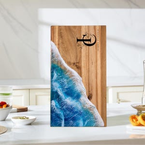 May include: Rectangular wooden chopping board featuring a blue and white resin wave design on the left, and natural wood on the right. The board is engraved with the letter "P", floral details, and the name "ISABEL CLAUDIA".