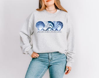 May include: A light gray sweatshirt featuring a design of blue and white seashells. The text "from shore to shore, His love endures" is printed below the seashells. The model is wearing blue jeans.