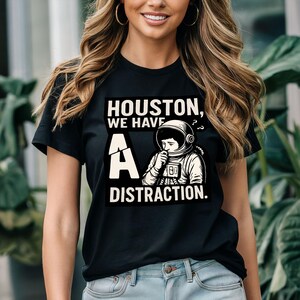 Puede incluir: Camiseta negra con el texto "Houston, We Have A Distraction". El gráfico presenta un astronauta con una expresión preocupada. La letra "A" se muestra de forma prominente, con la palabra "Distraction" debajo.