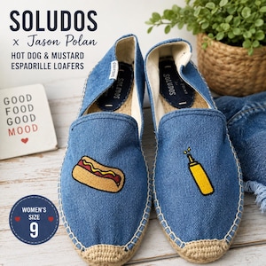 Puede incluir: Mocasines alpargatas de mezclilla azul con diseños bordados de un perrito caliente y una botella de mostaza. Los zapatos tienen una suela envuelta en yute y un interior de tela blanca. El texto dice SOLUDOS x Jason Polan HOT DOG & MUSTARD.