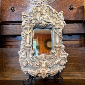 May include: A rectangular wall mirror featuring an ornate, light brown resin frame decorated with sculpted cherubs, floral motifs, and architectural columns. The central glass is surrounded by intricate relief carvings of winged figures.
