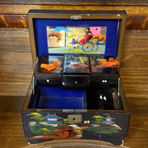 May include: A black lacquered wooden jewelry box with a hinged lid and blue velvet interior. The exterior features a painted landscape with pagodas. The interior lid has a mirror with a colorful graphic of a person in a rickshaw.