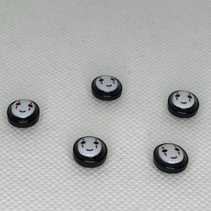May include: Five small, round black and white charms with a smiling face design. The charms have a black border and a white face with red accents. The charms are scattered on a white surface.