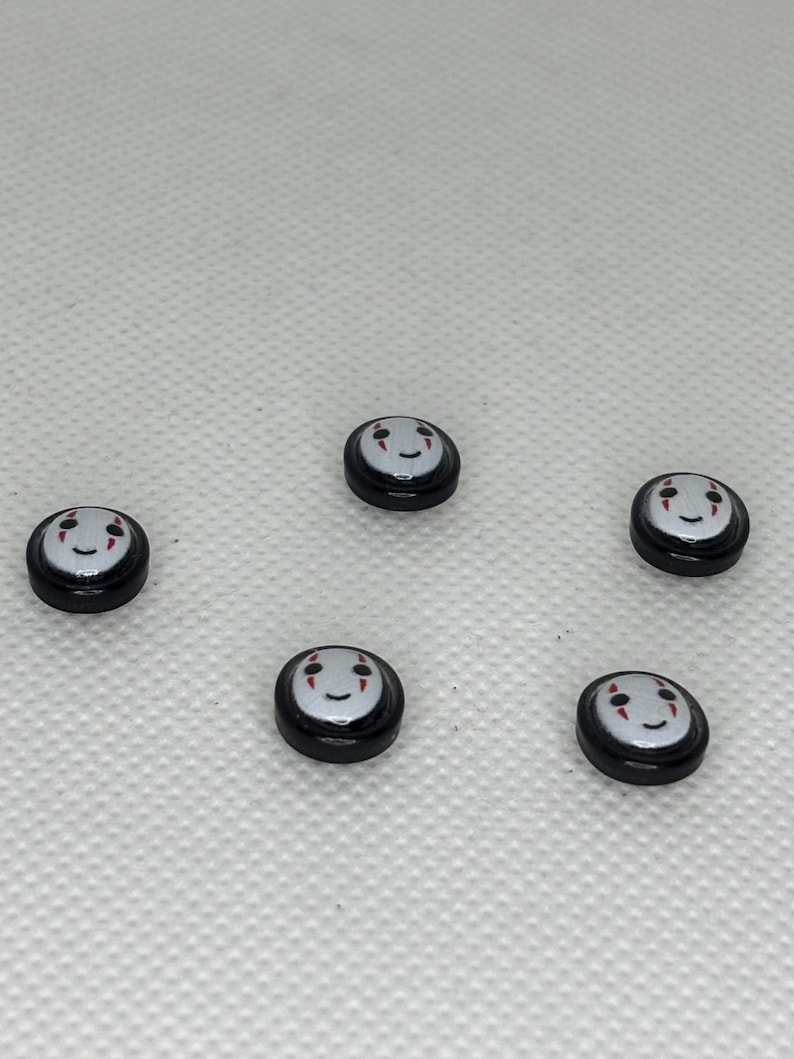 May include: Five small, round black and white charms with a smiling face design. The charms have a black border and a white face with red accents. The charms are scattered on a white surface.