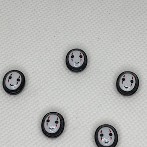 May include: Five small, round, black buttons with a white face design. The face has a smiling mouth, two red vertical lines for cheeks, and two black dots for eyes. The buttons are arranged in a circular pattern.