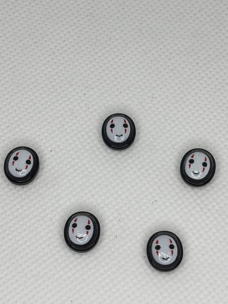 May include: Five small, round, black buttons with a white face design. The face has a smiling mouth, two red vertical lines for cheeks, and two black dots for eyes. The buttons are arranged in a circular pattern.