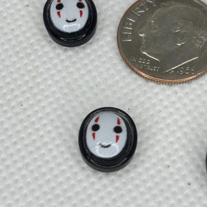 May include: Two small, round, black and white buttons with a cartoon face design. The face has black eyes, red cheek marks, and a smiling mouth. A U.S. dime is next to the buttons for size comparison.