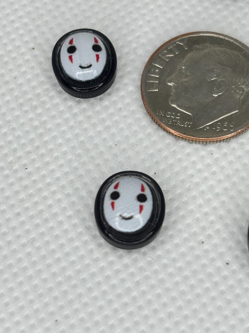 May include: Two small, round, black and white buttons with a cartoon face design. The face has black eyes, red cheek marks, and a smiling mouth. A U.S. dime is next to the buttons for size comparison.