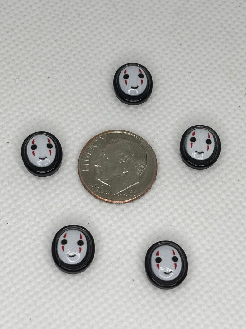 May include: Five small, round, black and white buttons with a smiling face design, arranged around a dime. The buttons have a gray background with red accents for the eyes and mouth. The dime is silver and shows a profile.