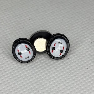 May include: Two black and white ear gauges featuring a smiling face design with red accents. The gauges are round with a black border and a white face. A black magnetic disc is behind the gauges.