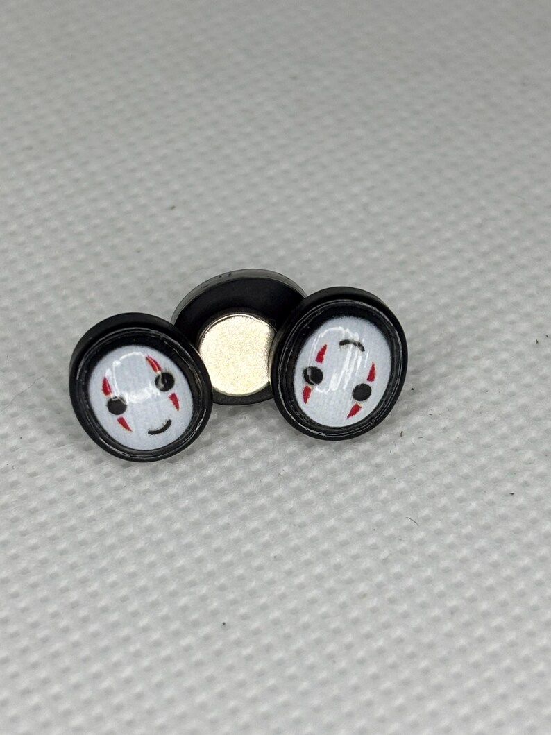 May include: Two black and white ear gauges featuring a smiling face design with red accents. The gauges are round with a black border and a white face. A black magnetic disc is behind the gauges.