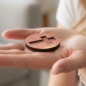 May include: A round, wooden pendant with a cross cutout. The pendant is a warm brown color and is held in an open hand. The cross is centered within the circle, creating a simple, yet elegant design. The pendant is likely a religious symbol.