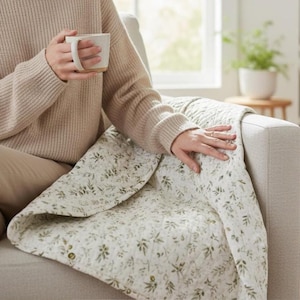 May include: A beige knit jumper and a white ceramic mug paired with a quilted throw blanket featuring a green botanical leaf pattern on a white background. The blanket is draped over a neutral-coloured sofa, showcasing detailed stitching.