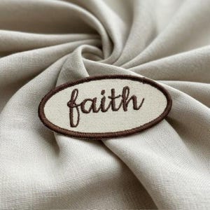 May include: An oval-shaped fabric patch featuring an off-white background with a dark brown embroidered border. The word faith is stitched in the center in a dark brown cursive script font, creating a clear contrast against the light fabric.
